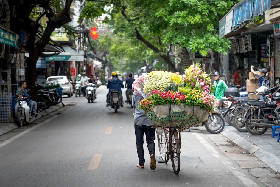 A street scene in West Hampstead featuring a person pushing a bicycle loaded with a large, colorful bouquet of fresh flowers wrapped in light-colored paper and tied with a ribbon. The bouquet includes