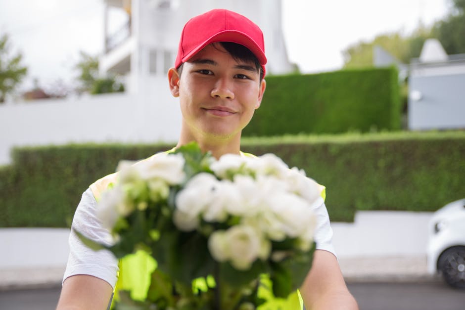 A young delivery person wearing a red cap and a light-colored shirt holds a bouquet of white roses and other white flowers wrapped in clear floral paper. He stands outdoors against a backdrop of green