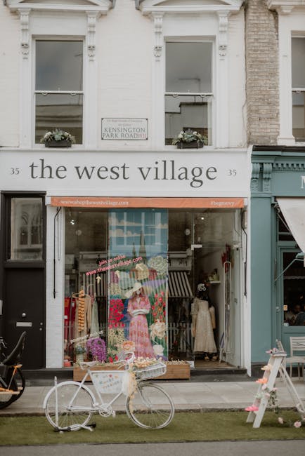 The storefront of 'the west village' on West End Lane West Hampstead, featuring a display window with colorful flower arrangements and a mannequin dressed in a pink floral dress inside. In front of th