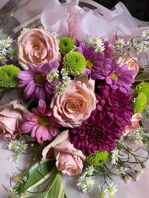 A vibrant floral bouquet featuring soft pink roses, purple daisies, bright green chrysanthemums, and white daisy-like flowers with yellow centers, all arranged with lush greenery and wrapped in delica