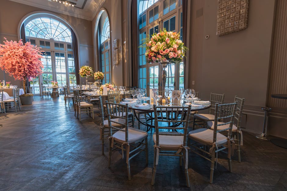 An elegantly arranged indoor event space featuring round dining tables adorned with tall, glass vases filled with lush floral arrangements of pink roses, white peonies, and green foliage. The tables a
