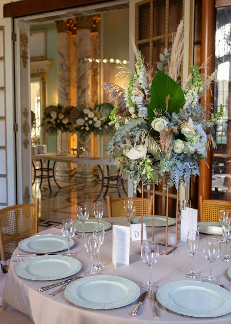 A floral arrangement featuring a mix of white roses, large green leaves, wispy pampas grass, and textured foliage, arranged on tall copper stands as a centerpiece on a round dining table. The bouquet