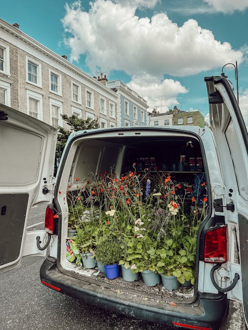 The open back of a delivery van filled with potted plants and freshly cut flowers, including red poppies, white daisies, and green foliage, arranged in light blue pots and scattered across the vehicle