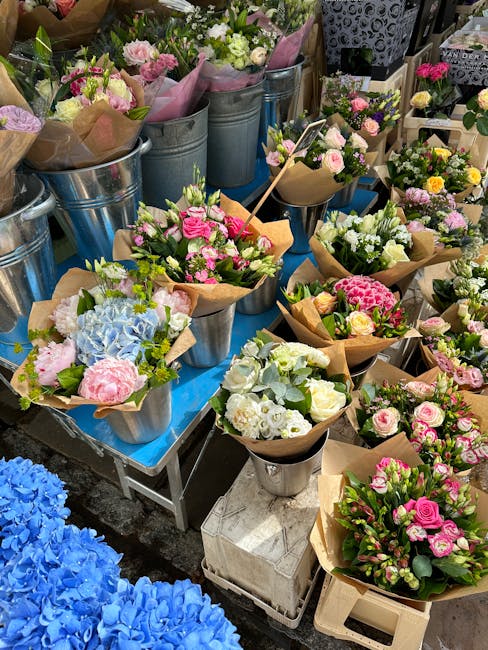An array of fresh floral bouquets arranged in metal buckets and brown kraft paper wrapping, featuring a variety of pastel-colored flowers including pink roses, white lisianthus, blue hydrangeas, and g