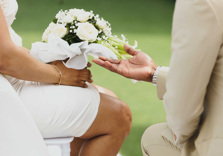 Close-up of a wedding bouquet featuring white roses, baby's breath, and greenery in white wrapping paper, being handed over by a person in white attire to a seated bride in a white dress, with a groom