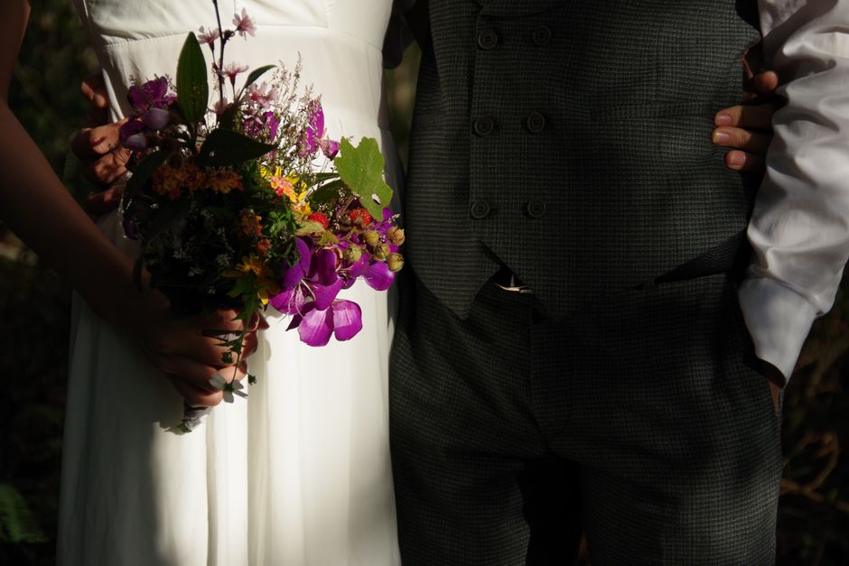 A bride holding a vibrant, mixed floral bouquet featuring purple orchids, yellow, pink, and purple blossoms, and green foliage, with petals and leaves appearing fresh and textured. She is dressed in a