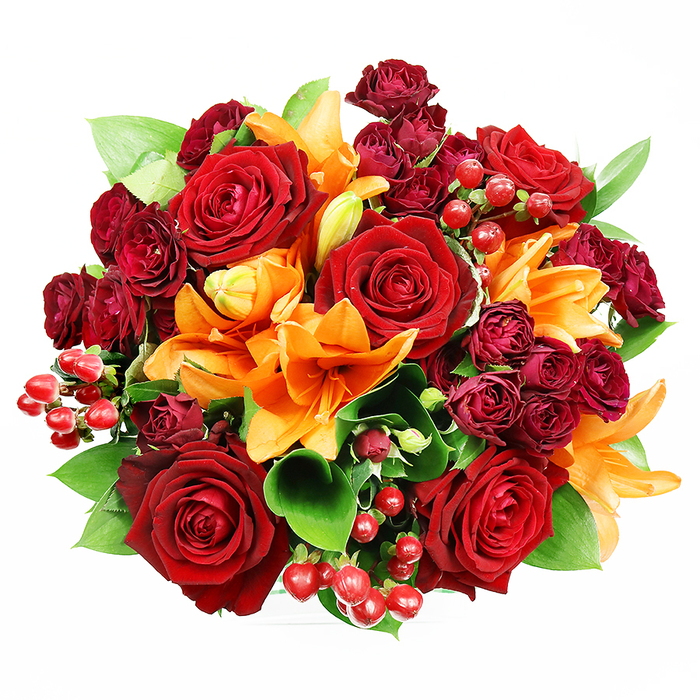 From a direct overhead view on a pristine white background, this densely packed round bouquet presents an opulent interplay of deep reds and fiery oranges softened by fresh green foliage. The composition is dominated by several fully opened deep red roses whose layers of velvet-like petals curl delicately into detailed centres, conveying a timeless sense of passion and warmth. Interspersed among those are radiant Asiatic lilies in a vivid orange hue-some flowers thrown wide to reveal their stamens, others partially unfurled or still in bud-bringing movement and sculptural form to the circle. Around the core, clusters of small, dark red spray roses add concentrated jewel-toned texture, while glossy hypericum berries scatter bright red highlights that reflect light and add playful punctuation. Broad, lustrous leaves-likely salal or ruscus-emerge between blooms to give the design breathing space and a fresh, natural framework. The soft, even lighting highlights the contrast between the velvet of rose petals and the satiny sheen of lily segments, emphasising tactile differences you can almost feel. Scent memory is present in thought: the deep, familiar perfume of roses balanced by a faint citrus-spicy edge from the lilies. This bouquet reads as an elegant statement for romantic gestures, autumnal celebrations or special anniversaries and would suit a delivery to a West Hampstead home or a thoughtful gift collected near Mill Lane or the local market-crafted with the same care a neighbourhood florist uses when dressing an occasion.